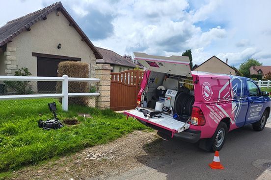 Intervention de debouchage dans une zone pavillonnaire. Le pick-up est arrêté devant la maison avec des plots de signalisation orange. Du matériel professionnel est sorti devant le véhicule